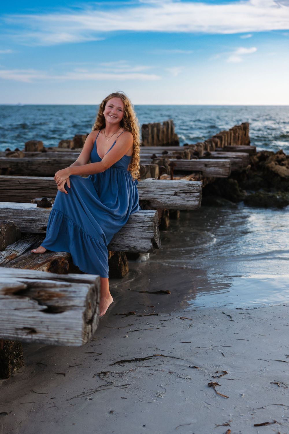 A woman in a flowy blue dress poses gracefully on a rocky beach during sunset, creating artistic seaside portraits.