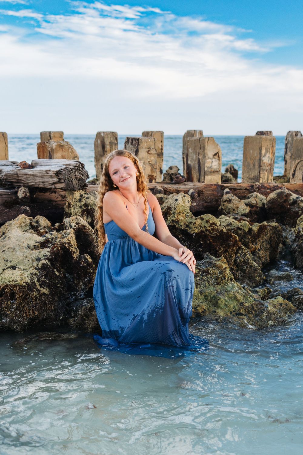 A figure in a blue flowing dress poses gracefully on rocky coastal outcrops as waves crash around.