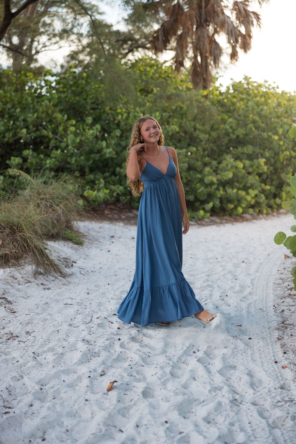 A blue maxi dress flows elegantly against white sand and tropical foliage at sunset.