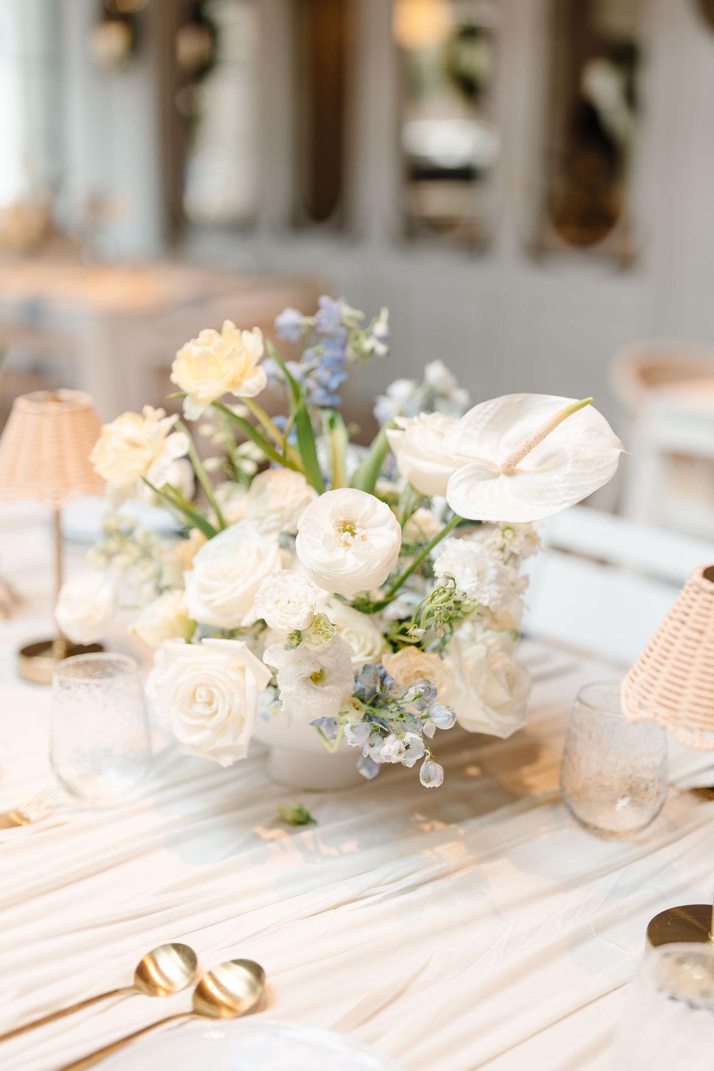 Elegant white floral centerpiece with roses and blue accents on a white tablecloth at a wedding reception.