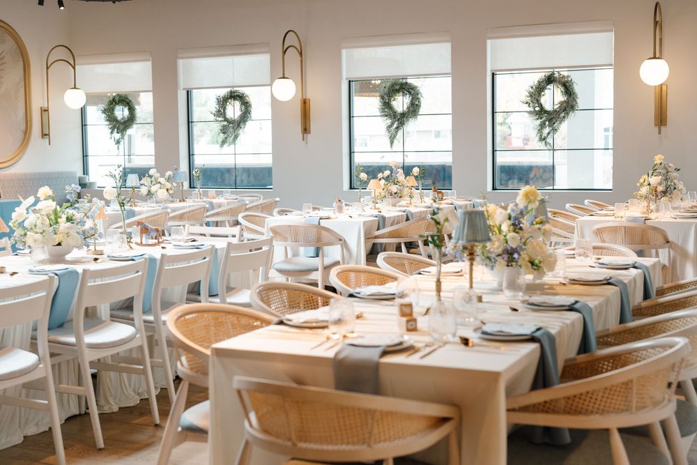 Bright wedding reception hall with wooden chairs, long tables, blue runners, and wreath-adorned windows.