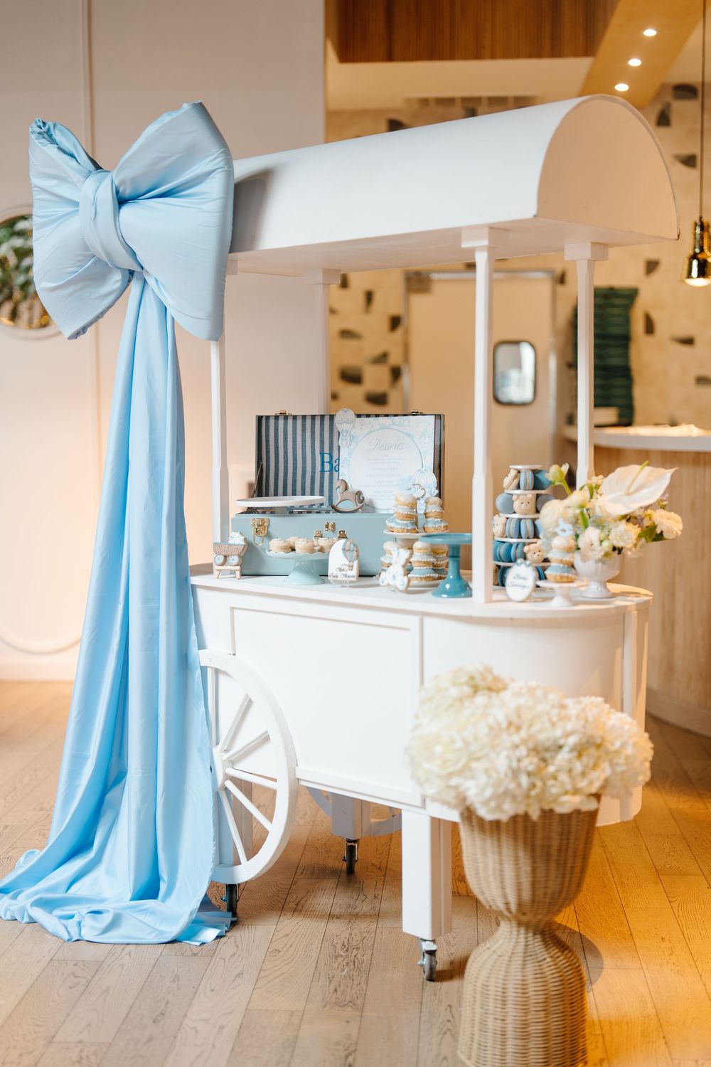 White dessert cart decorated with a large blue bow and macarons at a baby shower event.