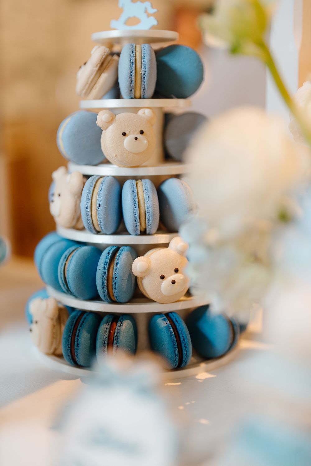 Tiered display of blue and white macarons with teddy bear decorations at a baby shower.