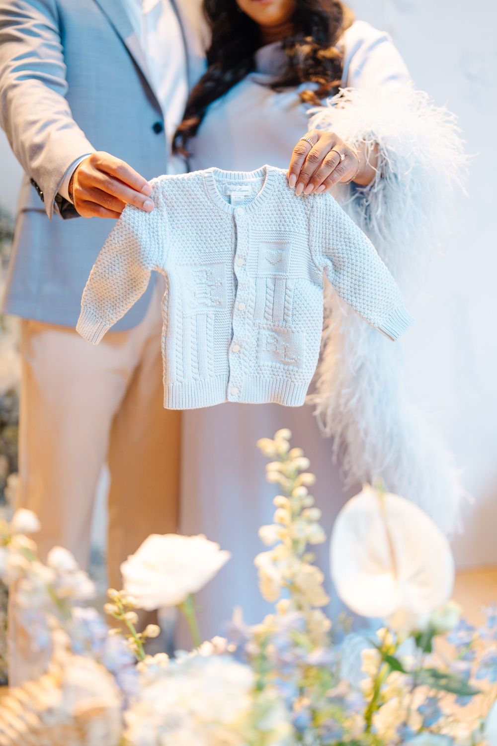 Couple holding a tiny white knit baby cardigan at a floral-decorated baby shower celebration.