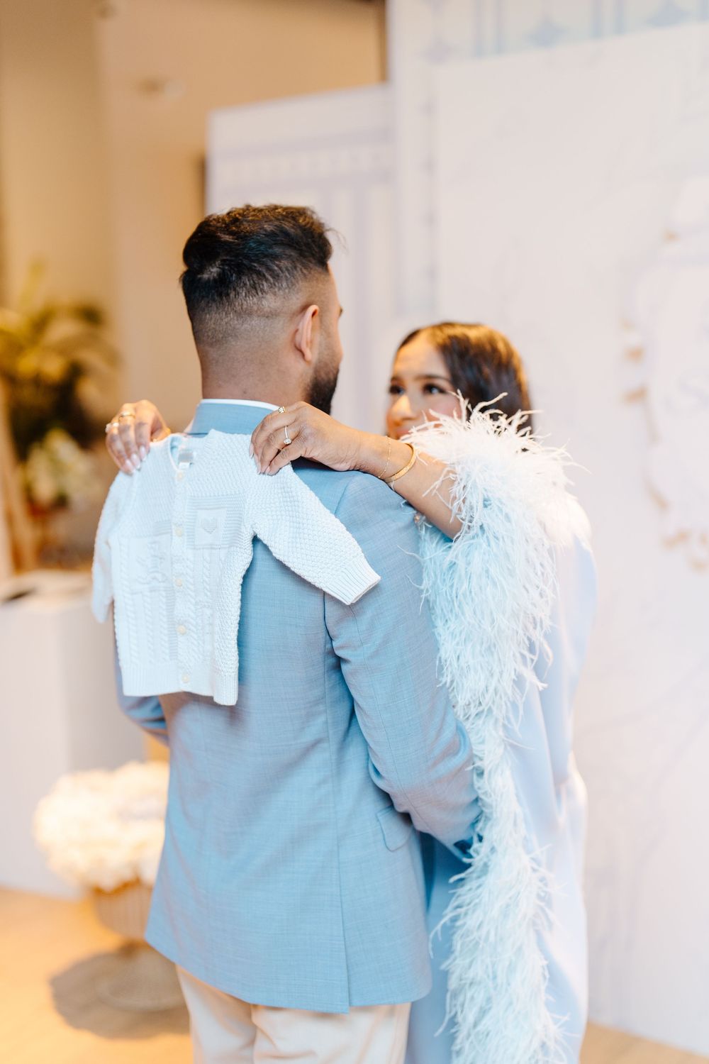 Couple embracing at baby shower, woman in blue feather wrap holding tiny white baby outfit.