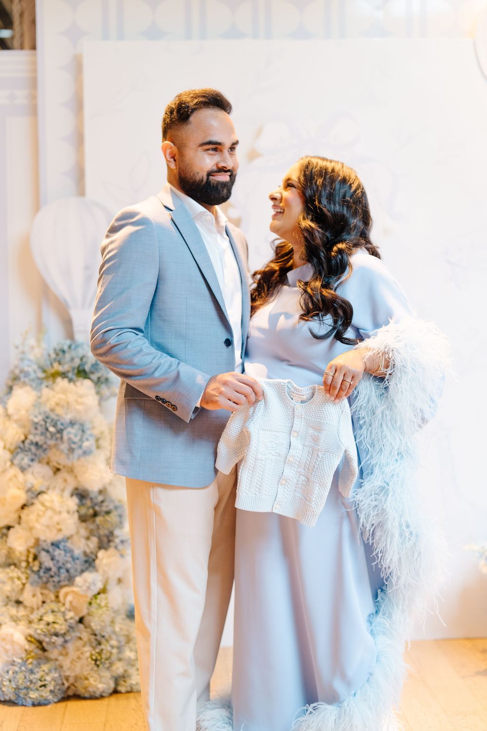 Smiling couple at baby shower holding white knit baby clothes, dressed in elegant blue outfits.