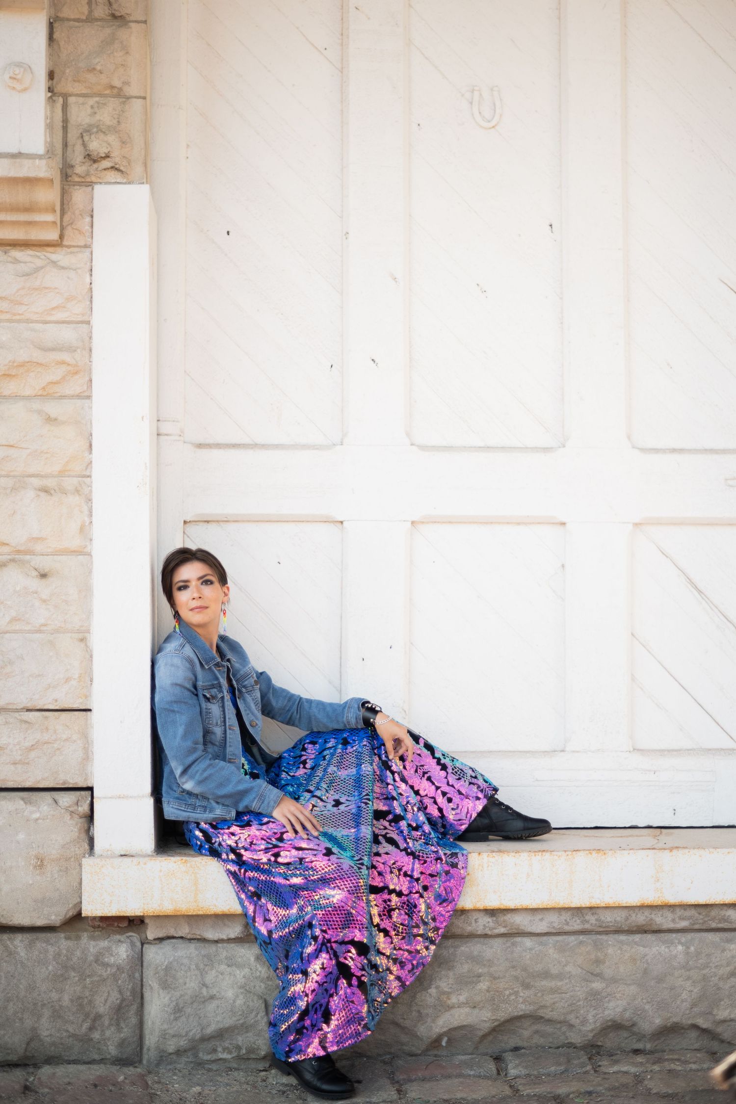 Person in denim jacket and vibrant purple floral skirt sits on stone steps against white building wall.