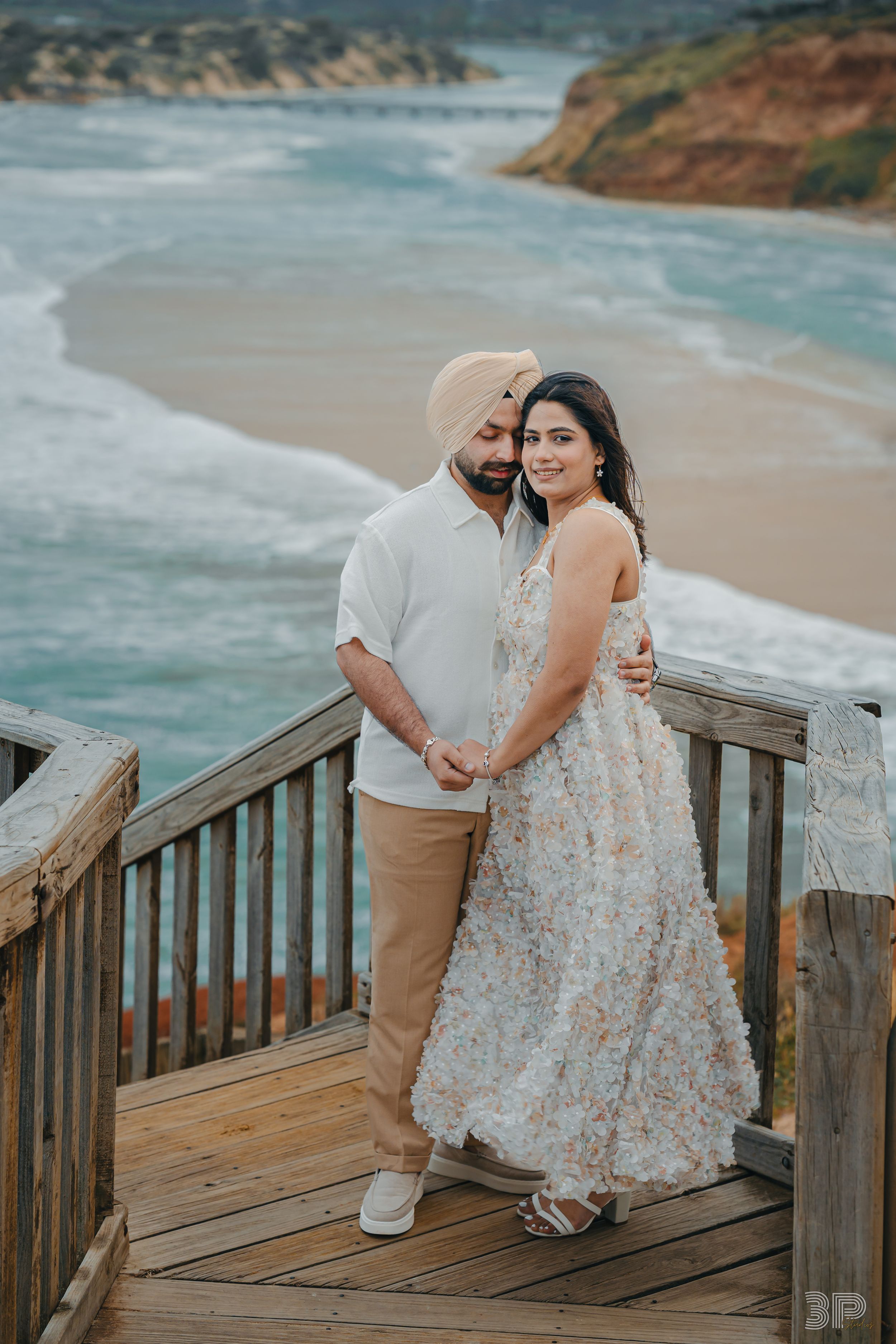A romantic couple poses on a wooden boardwalk overlooking a scenic beach with turquoise waters and cliffs.