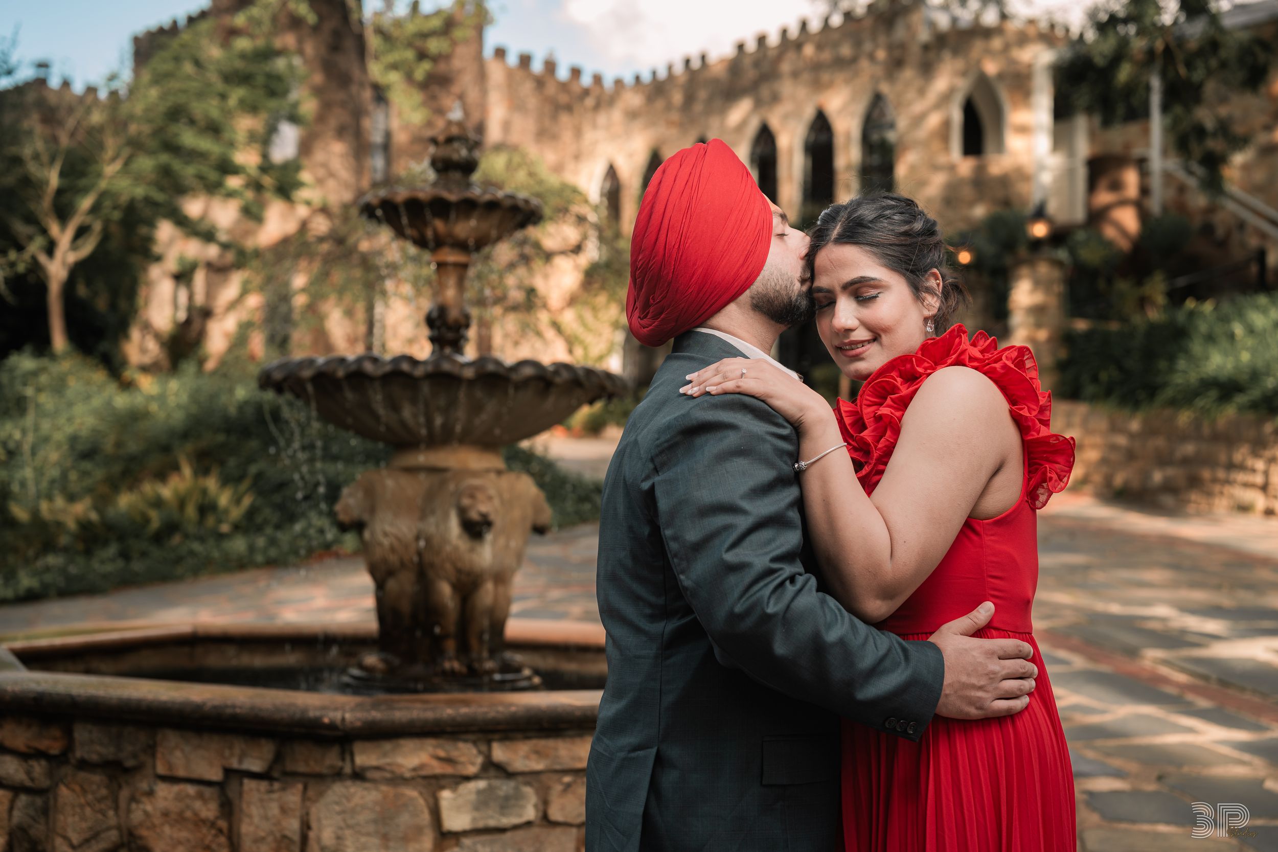 A couple in red and black attire embraces in a romantic outdoor photoshoot at a historic stone wall location.