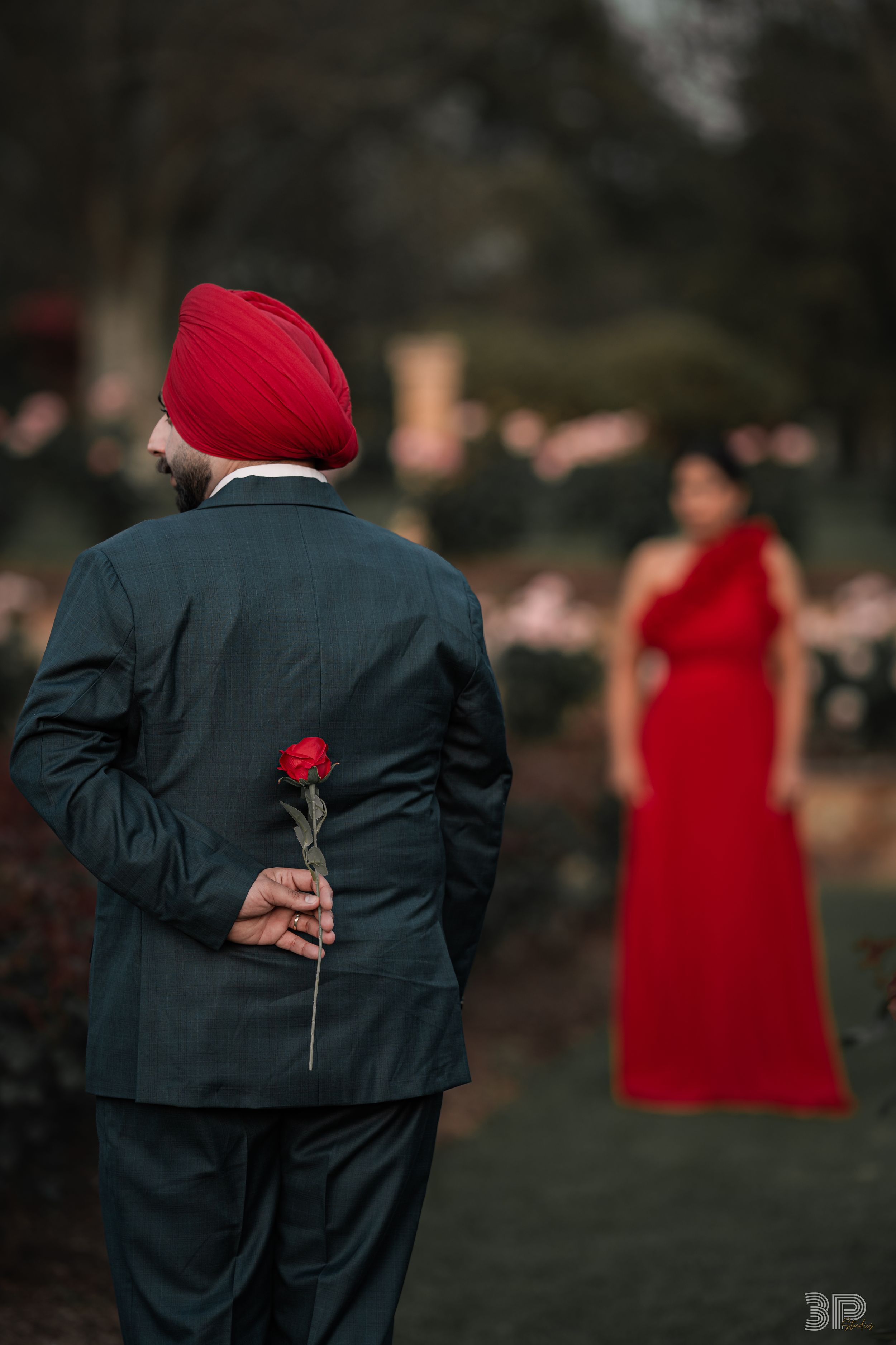 A person in a dark suit holds a red rose behind their back while someone in red stands in the distance.