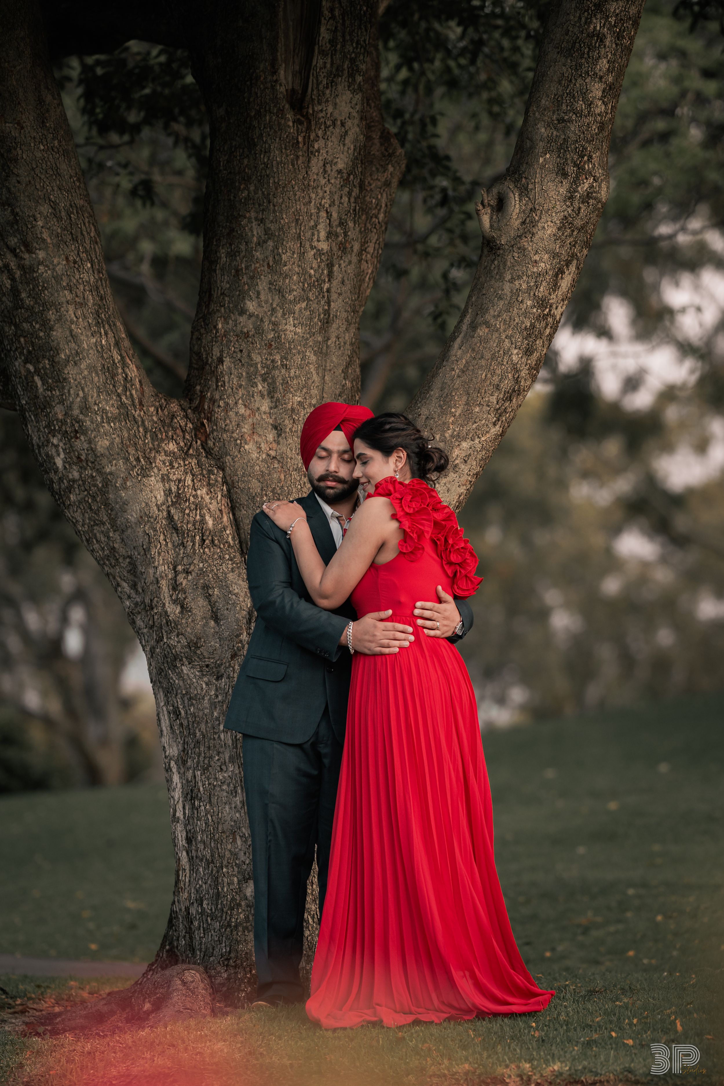 A romantic couple embraces beneath a tree, with one wearing a flowing red dress and the other in a dark suit.
