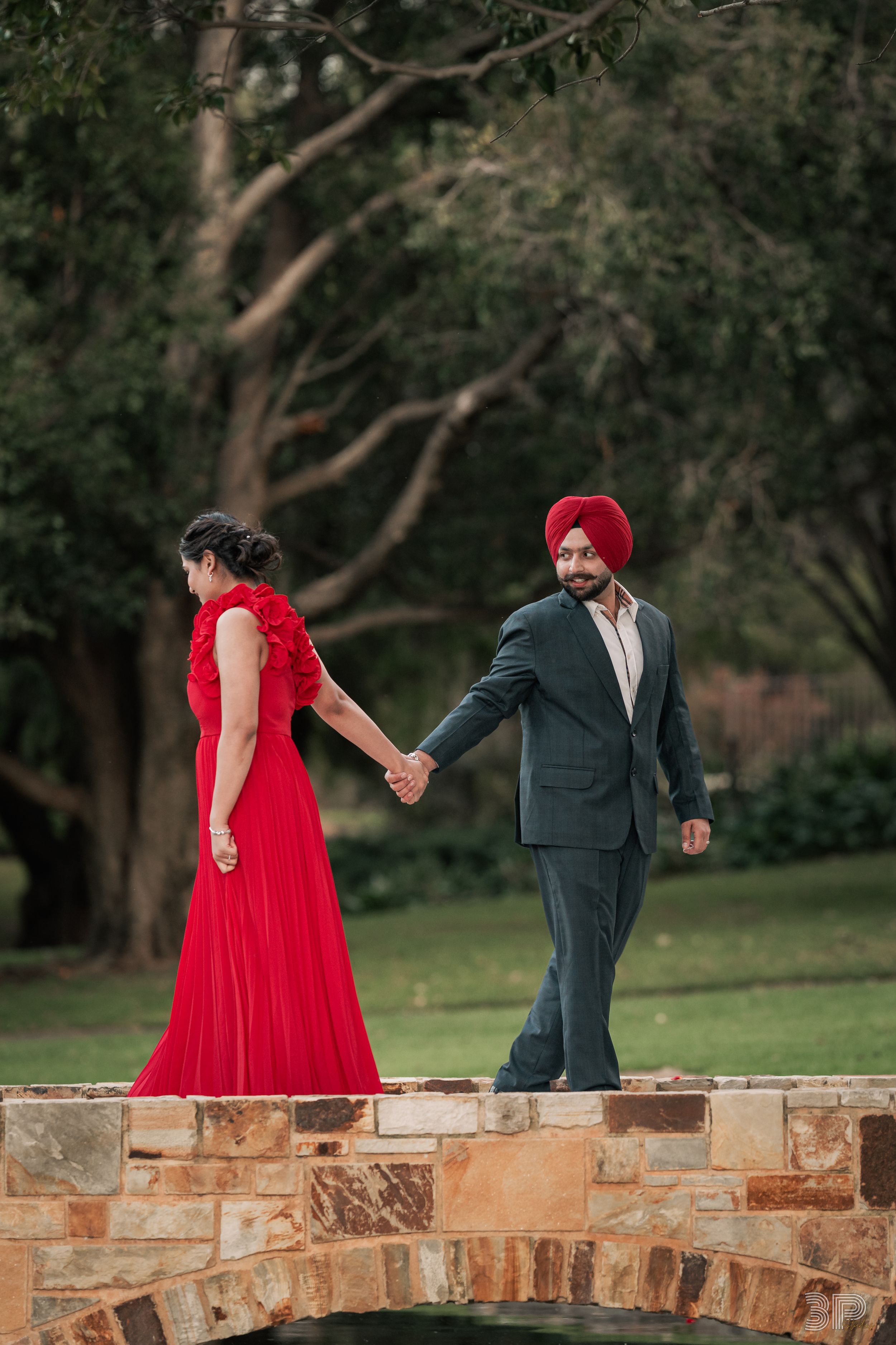 A couple in formal attire walks playfully along a stone wall surrounded by trees at dusk.