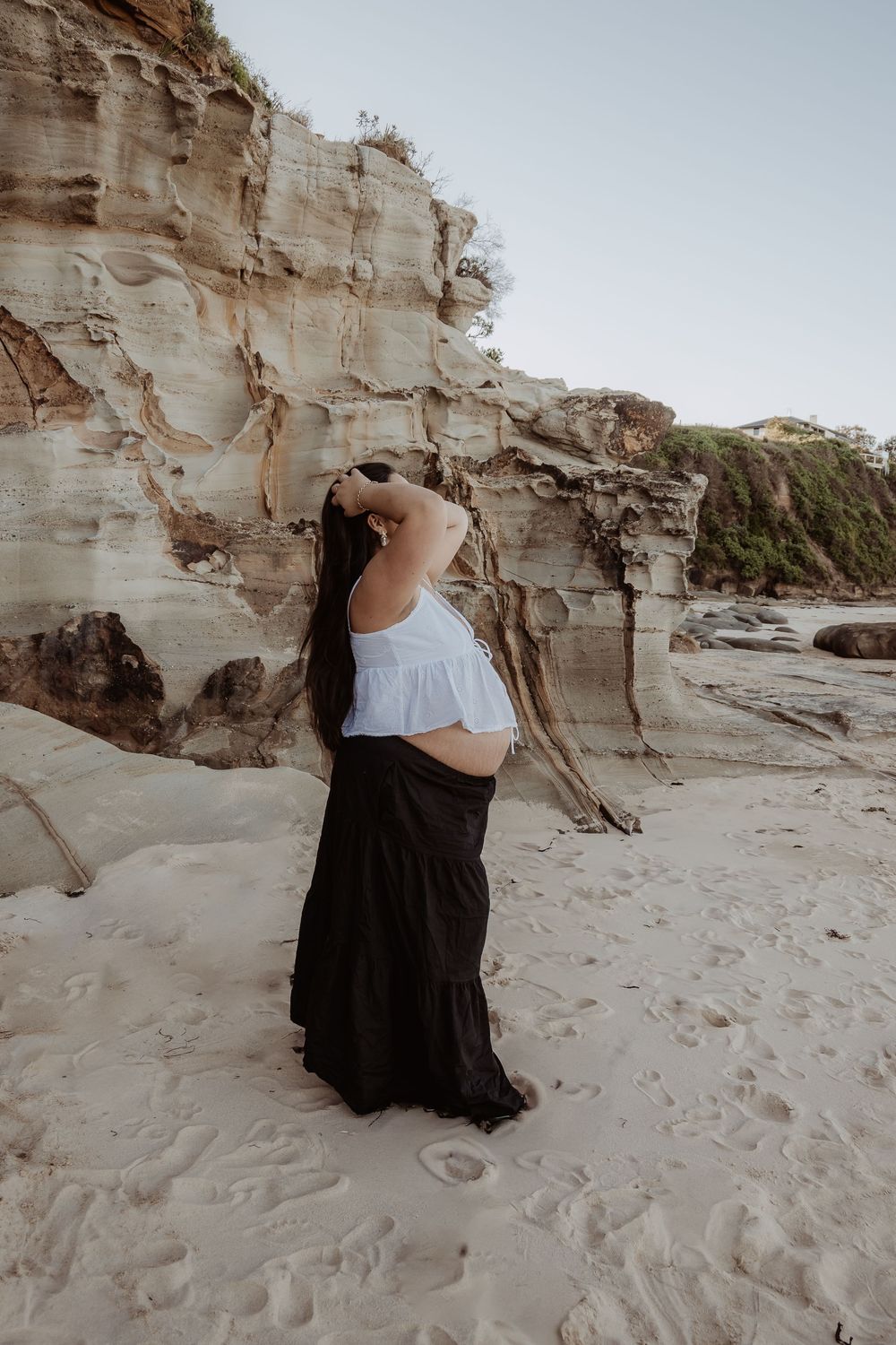 Pregnant woman in a white top and black skirt poses near coastal rock formations on a sandy beach at a central coast maternity photoshoot.