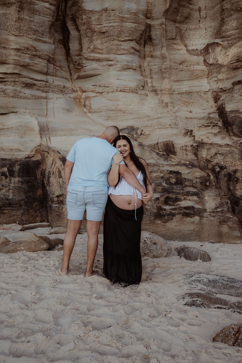 A couple poses together on a sandy beach against dramatic rocky cliffs during a central coast maternity photoshoot.