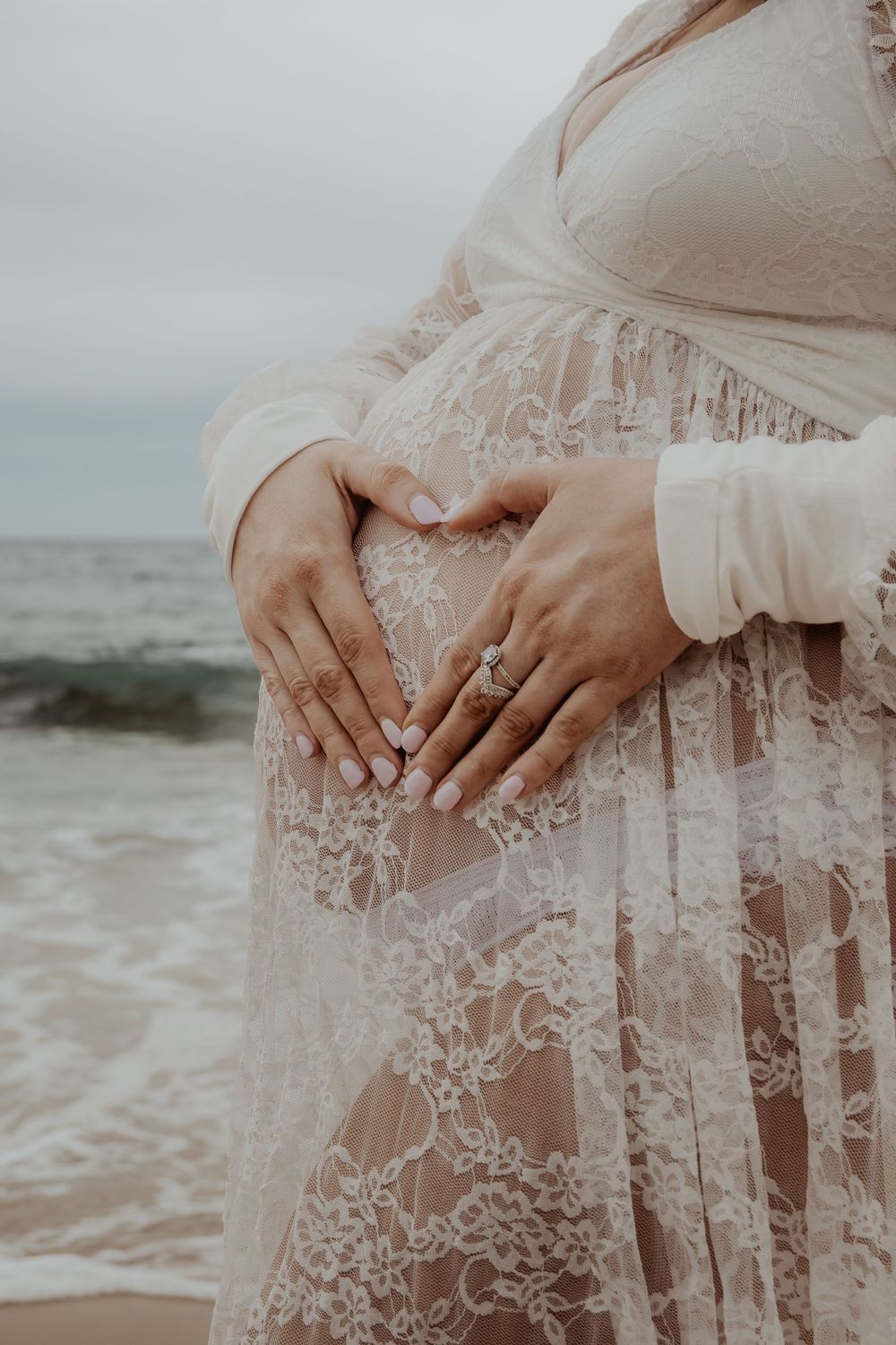 Close-up of delicate lace details and hands making a heart on a white dress by the ocean at a central coast maternity photoshoot.