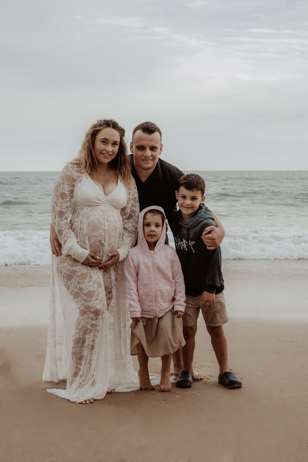 Family portrait on a sandy beach with ocean waves in the background during a central coast maternity photoshoot.
