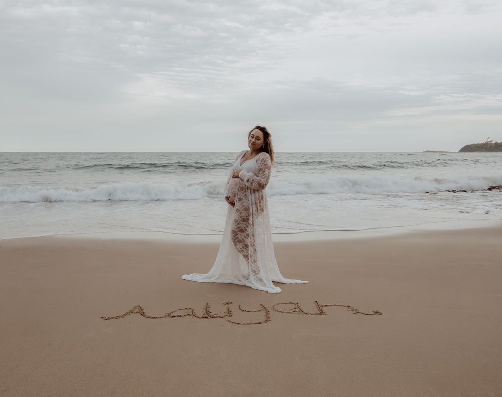 Pregnant woman in a flowing white lace dress stands on a beach with waves in the background during a central coast maternity photoshoot.