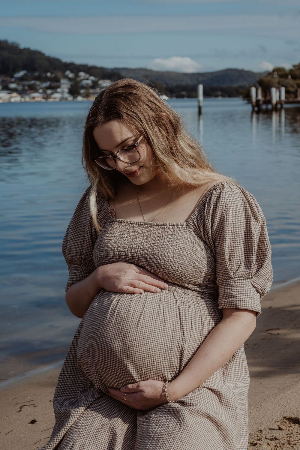Pregnant woman in brown dress poses by waterfront during golden hour during a central coast maternity photoshoot.