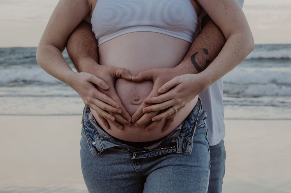 Hands forming heart shape over pregnant belly on beach at sunset during a central coast maternity photoshoot.