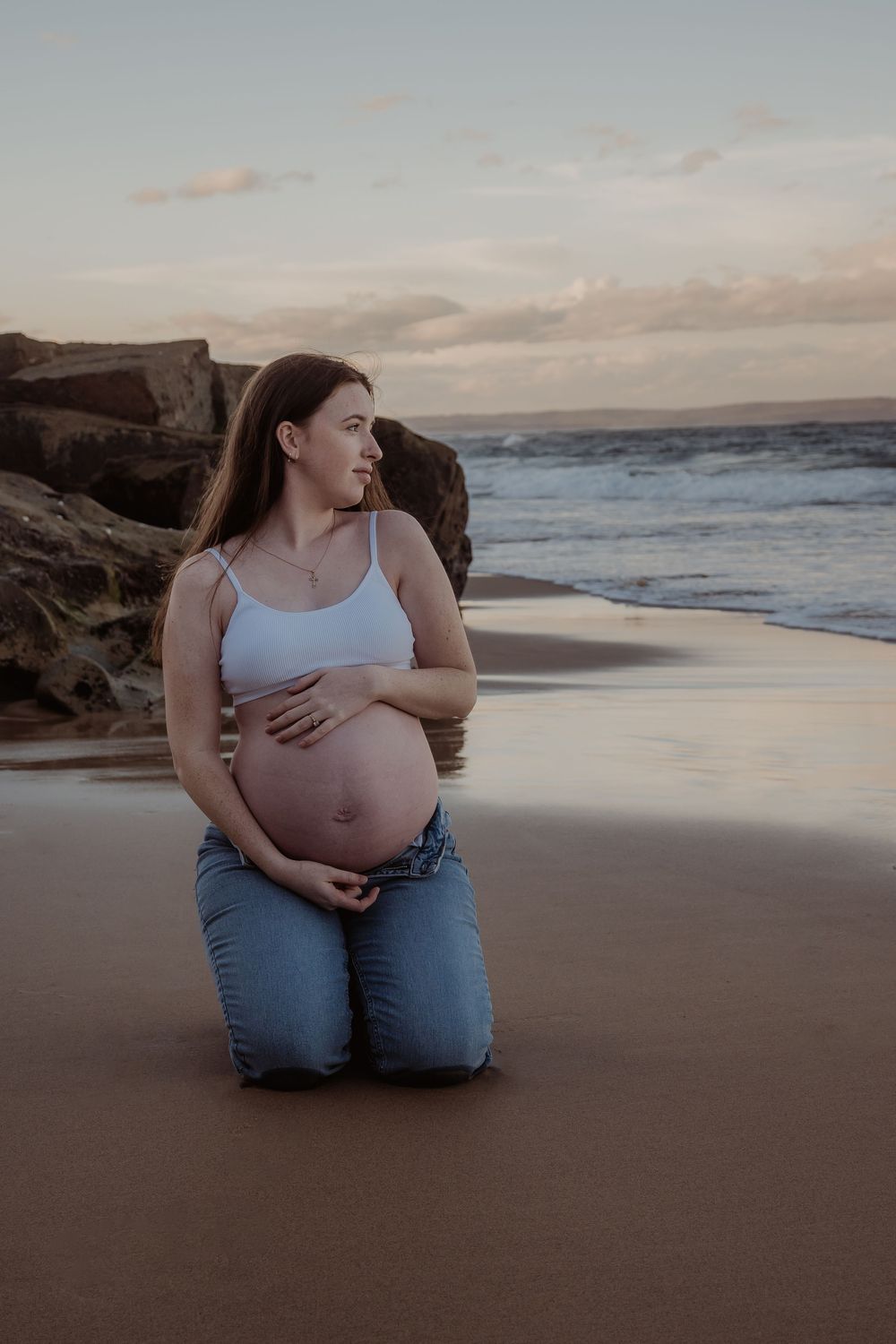 Pregnant woman embracing her belly sitting on the sand with water and rocks in the background during a central coast maternity photoshoot.