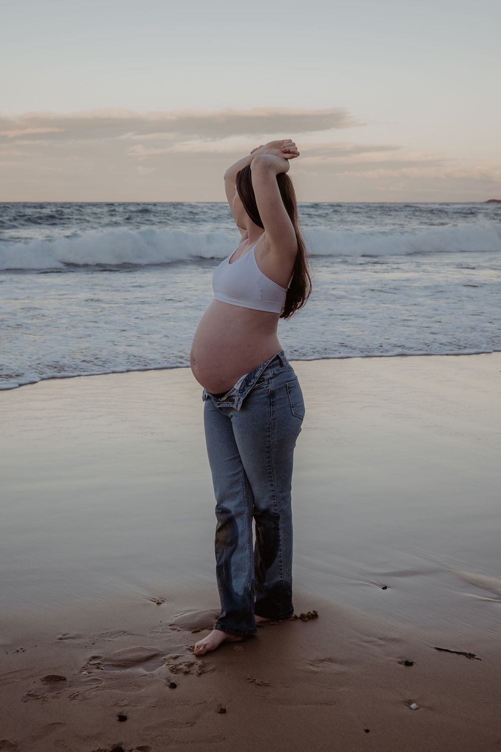 Pregnant woman in white top and jeans standing on beach at sunset with ocean waves in background at a central coast maternity photoshoot.