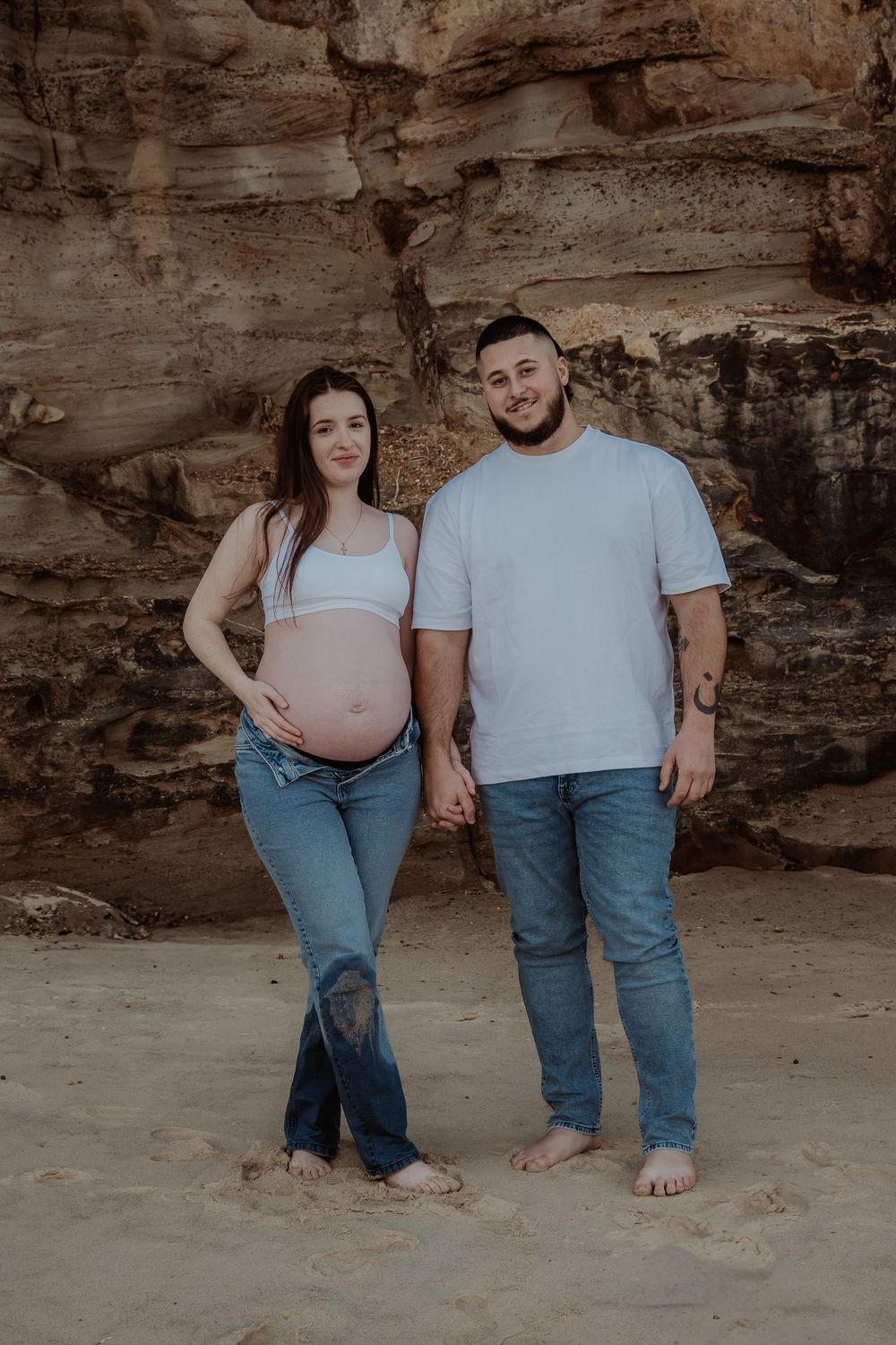 A couple stands barefoot on sandy beach against rocky cliff wall in matching denim and white outfits during central coast maternity session.