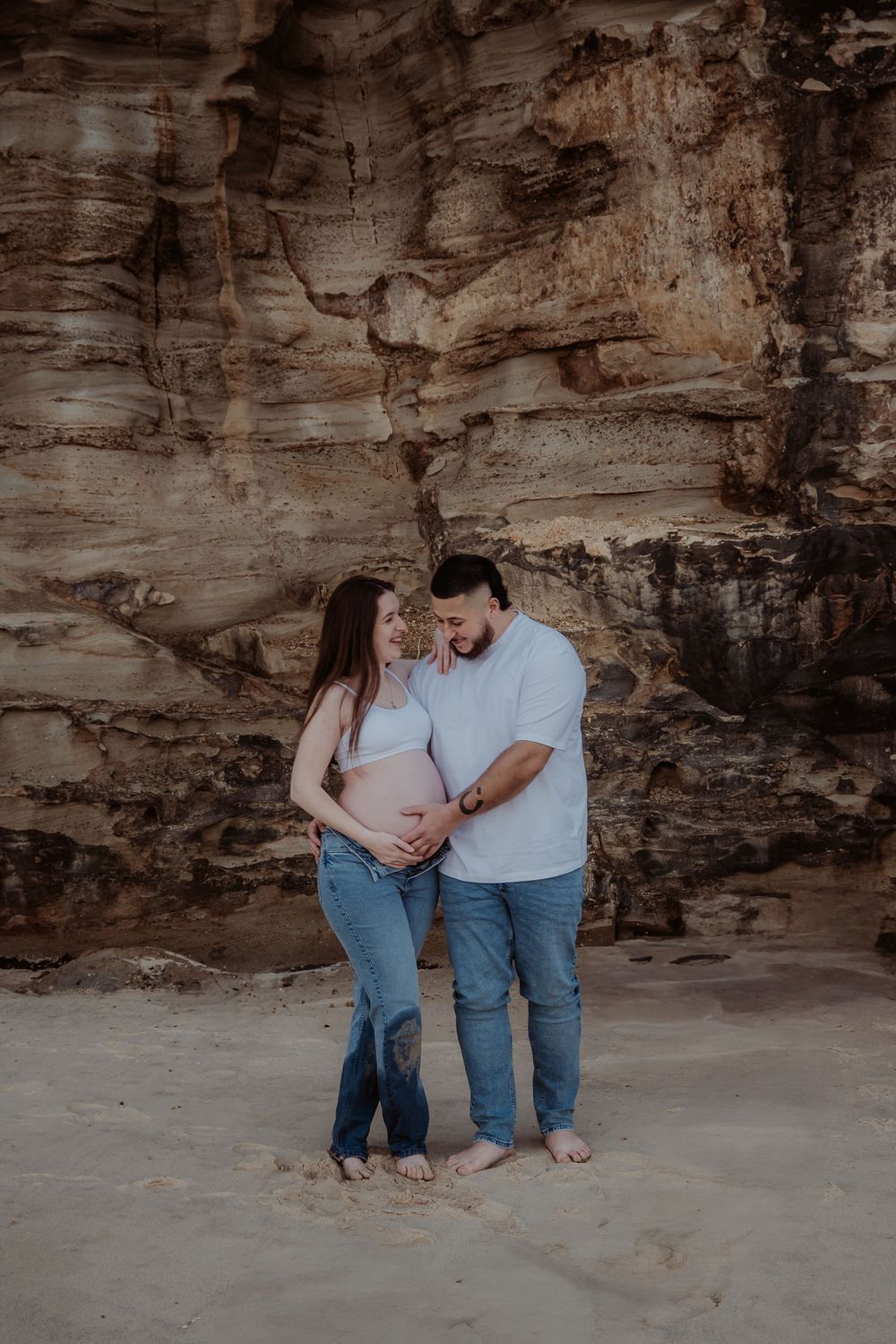A loving couple shares a tender moment against a rustic stone wall backdrop by the ocean at a central coast maternity photoshoot.
