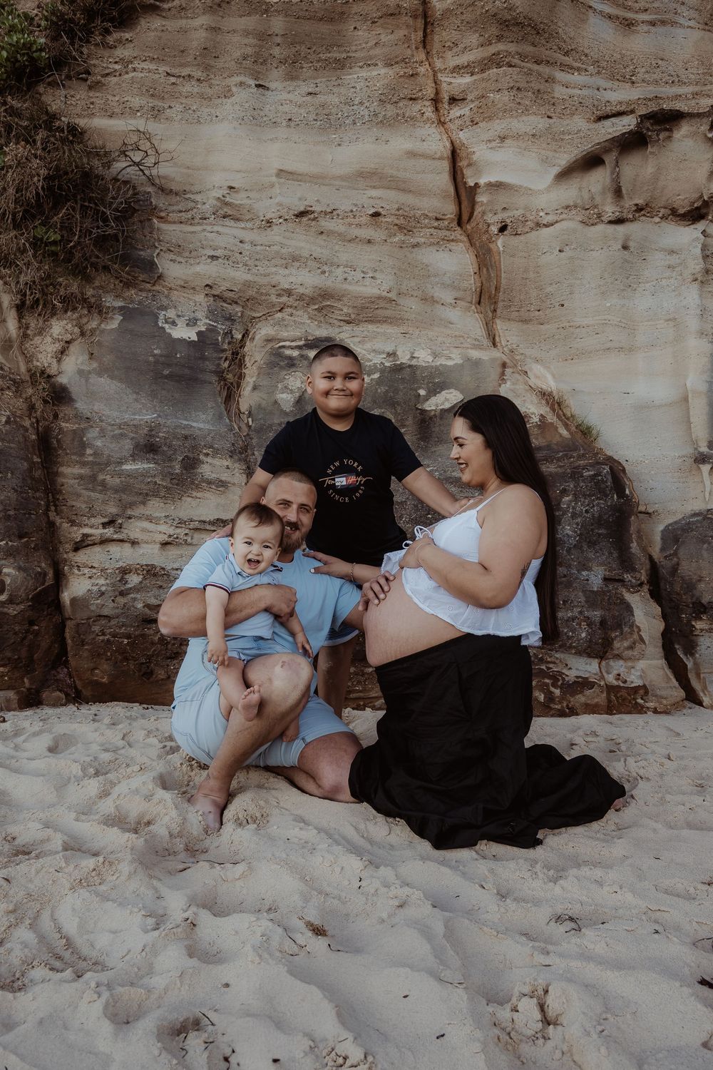 Family sitting together on sandy beach against rocky cliff backdrop during a central coast maternity session.