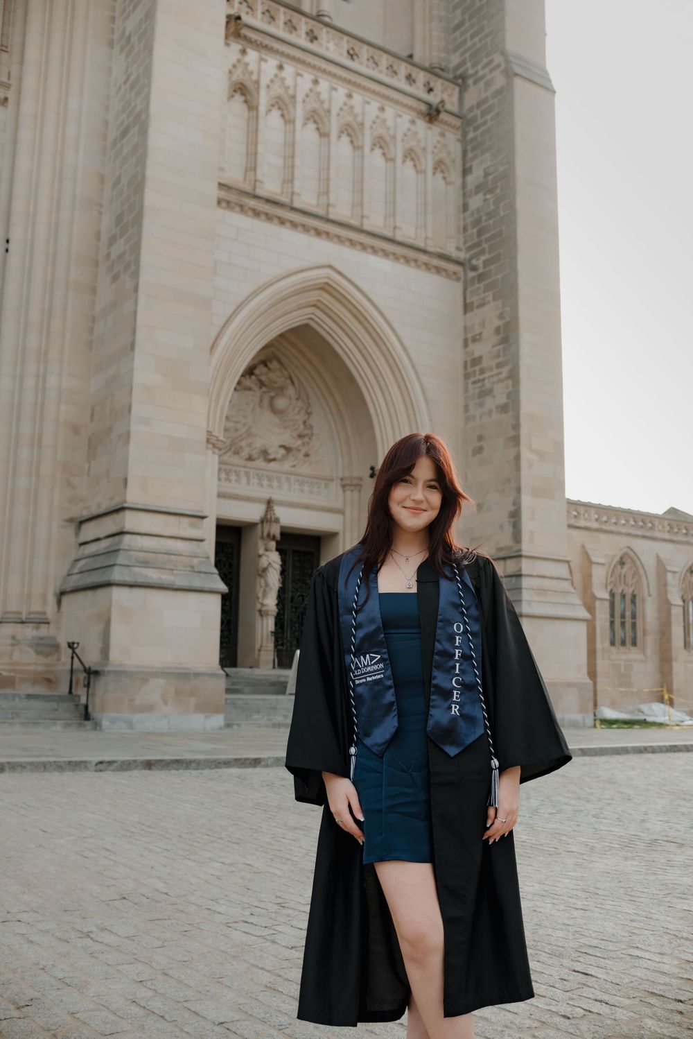 Graduate in black academic regalia and blue vest poses in front of gothic stone archway during graduation photoshoot.