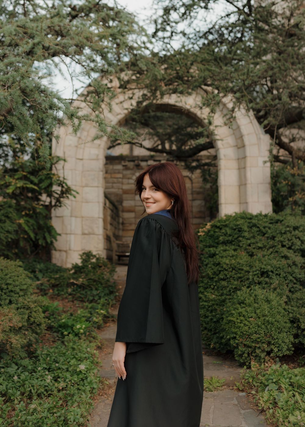 A figure in dark clothing stands against stone architecture surrounded by lush garden foliage.
