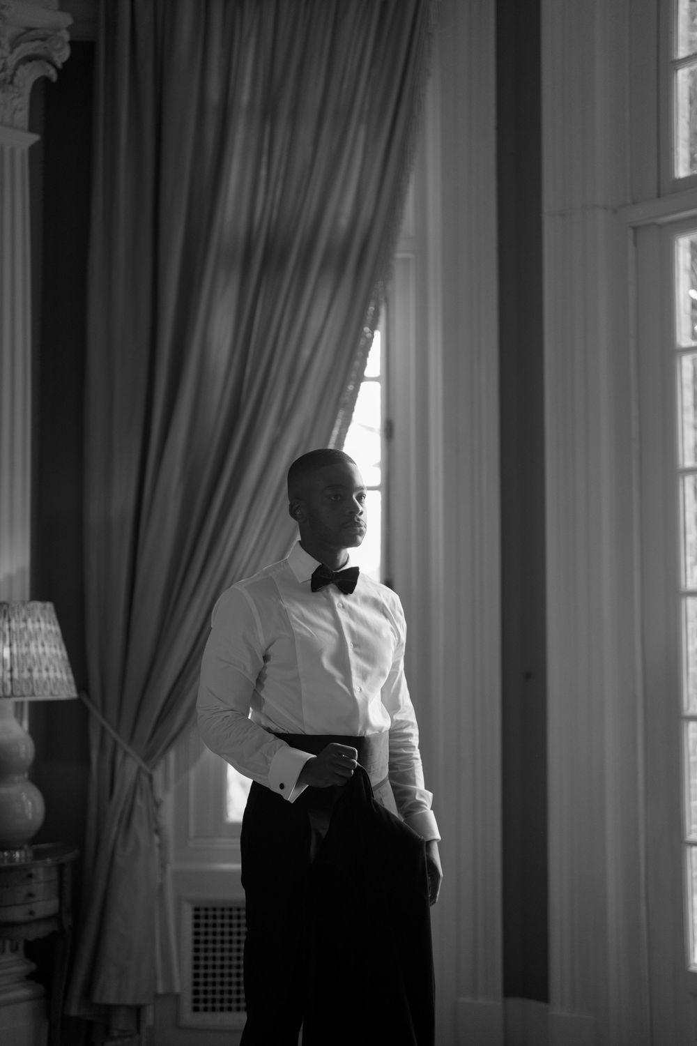 Dramatic black and white photo of figures standing by tall windows with flowing curtains.