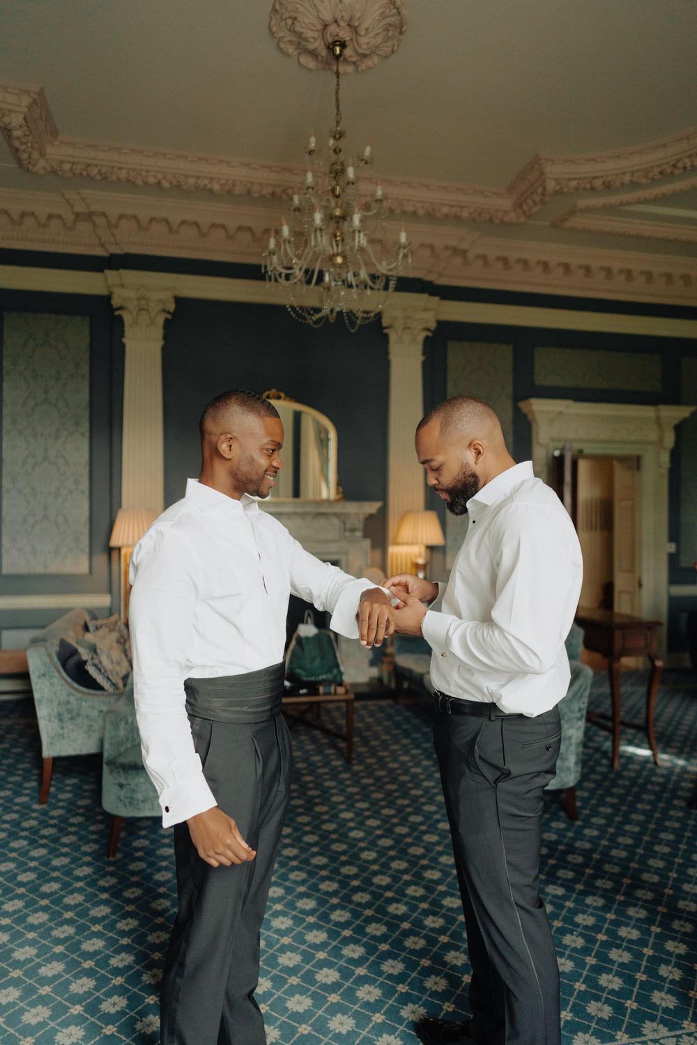 Groomsmen in white shirts and dark pants practice dance moves in an ornate ballroom with chandeliers.
