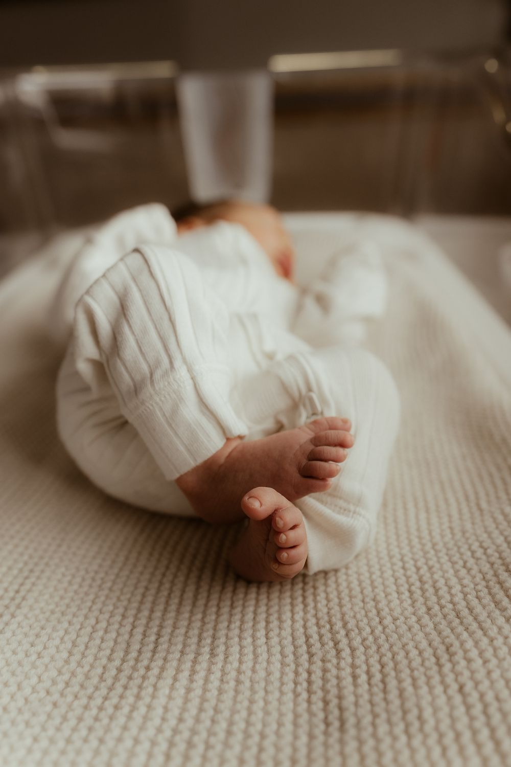 Small baby feet in white clothing rest on a cream-colored knitted blanket in a minimalist setting.