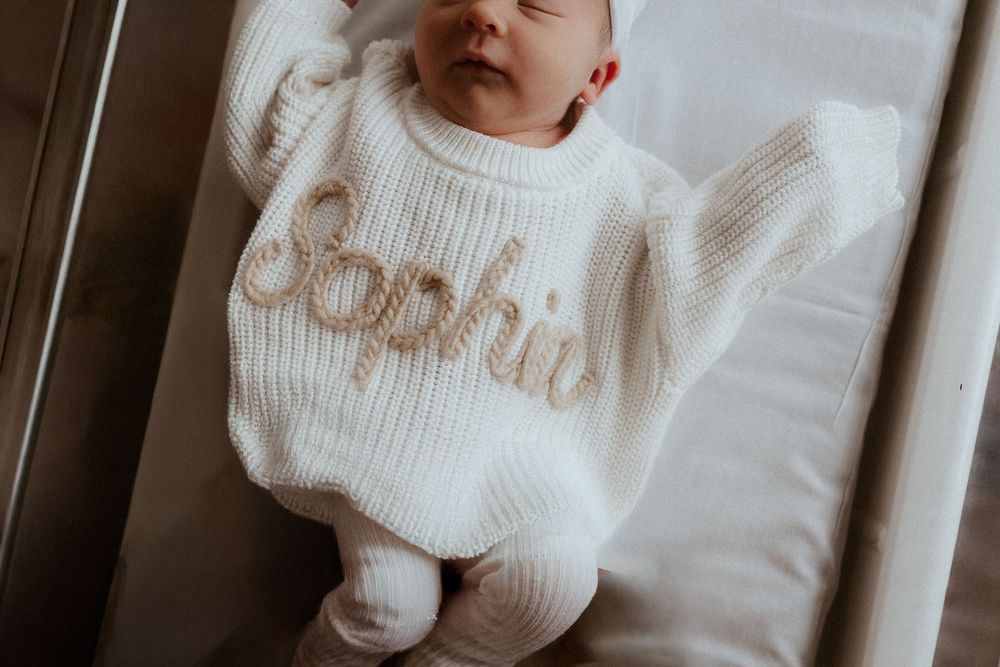 A newborn baby wearing a white knit sweater is held gently while resting.