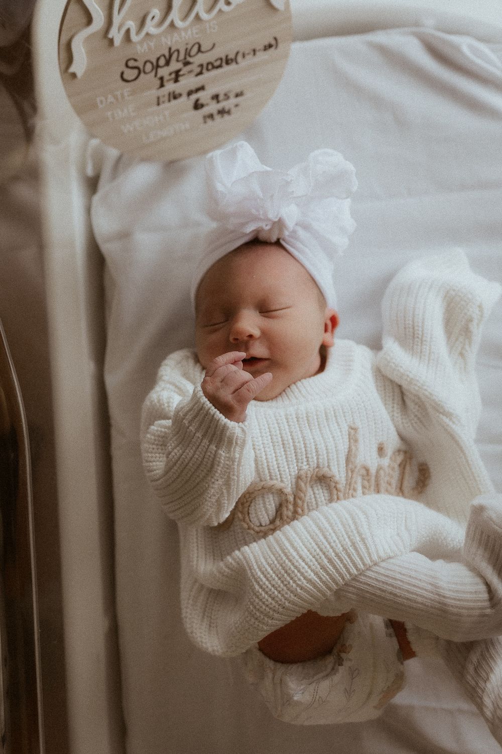 Newborn baby wearing white knit sweater sleeping peacefully on white bedding.