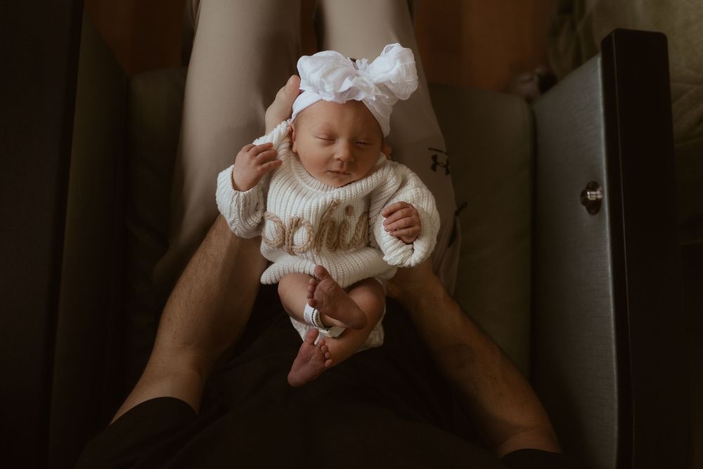 A baby wearing a white dress sits on a wooden floor while reaching for something out of frame.
