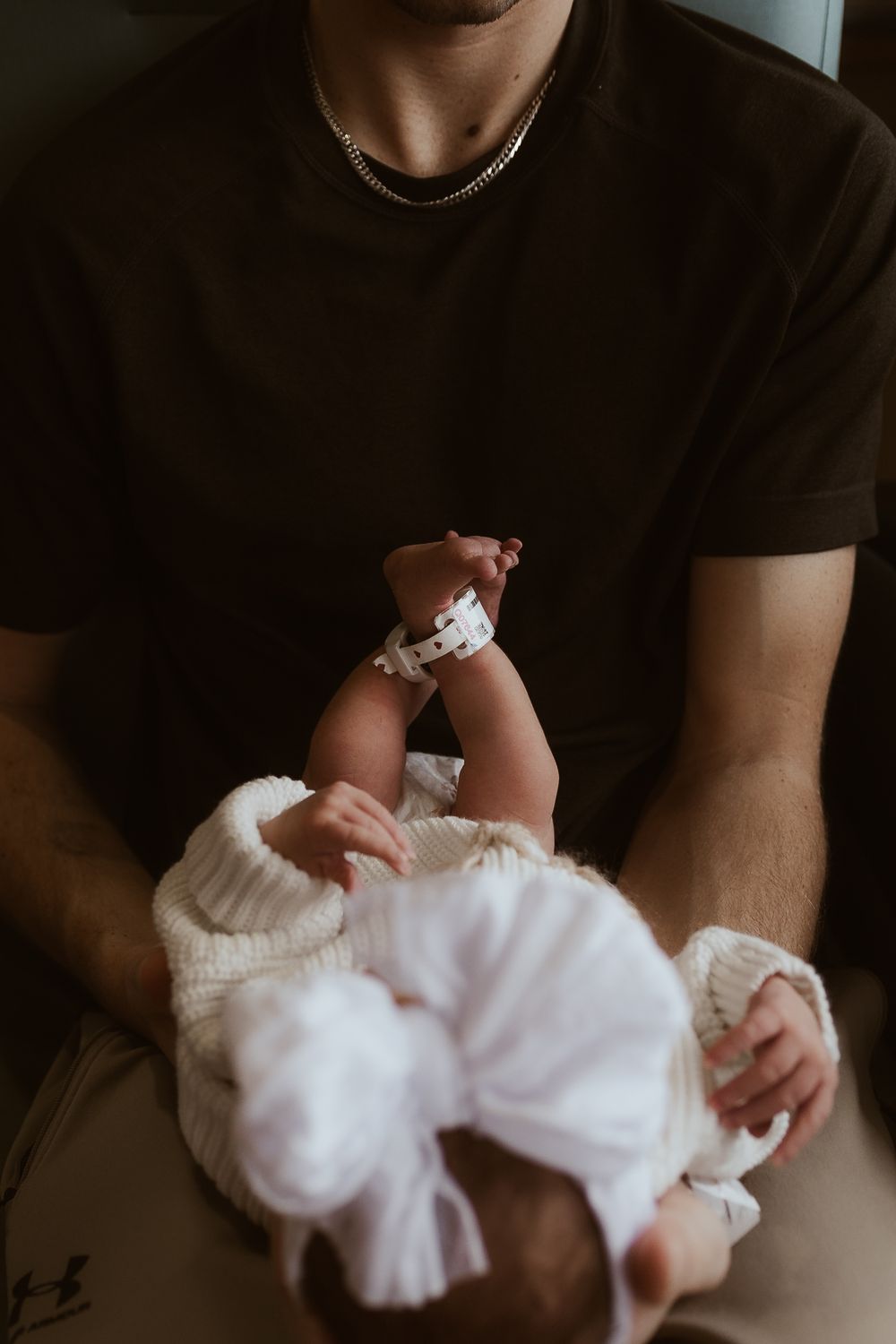 Baby in white outfit being cradled in parent's arms against dark background.