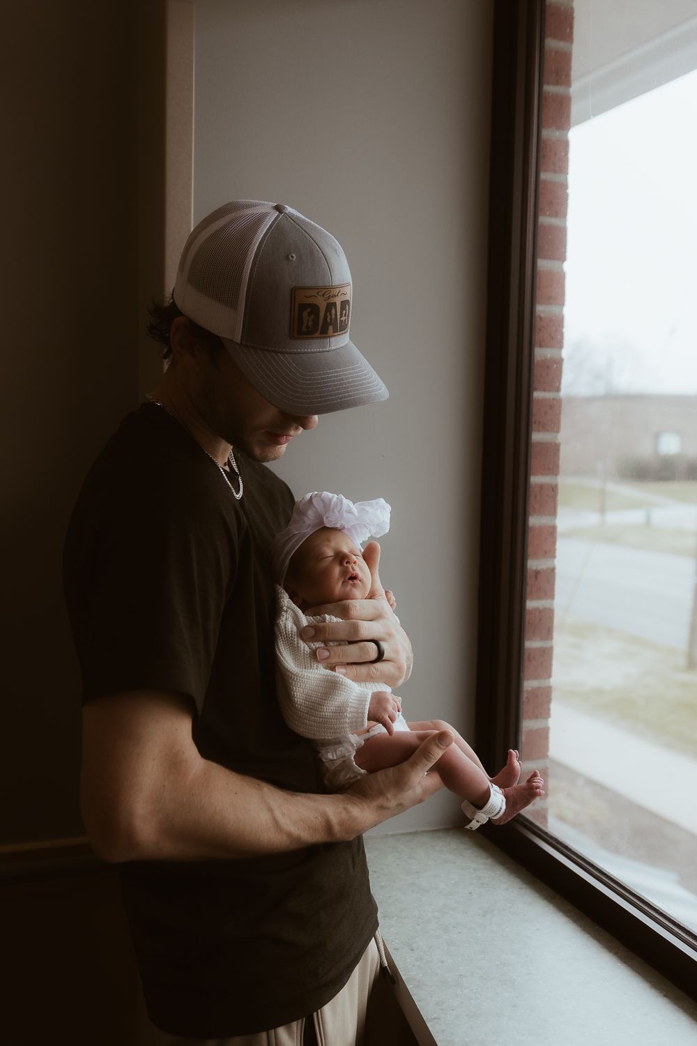 Parent in baseball cap holding infant while standing by window in moody lighting.