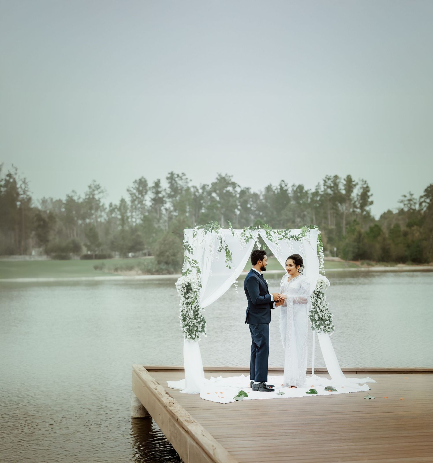 A romantic lakeside wedding ceremony setup with white decor and floral arrangements on a wooden dock.