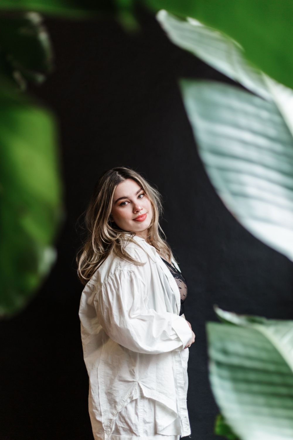 Fashion portrait with model in white ruffled blouse against green foliage and dark background.