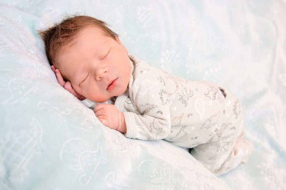 Newborn baby sleeping peacefully in soft white swaddle blanket on light blue background.