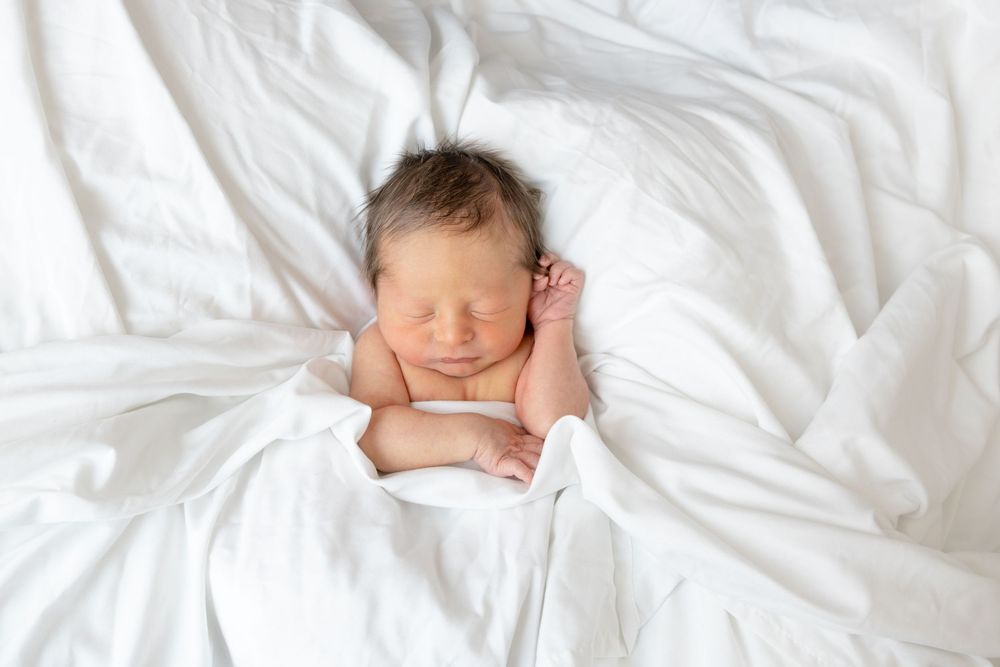 A sleeping newborn baby lies on white bedding with soft natural lighting.