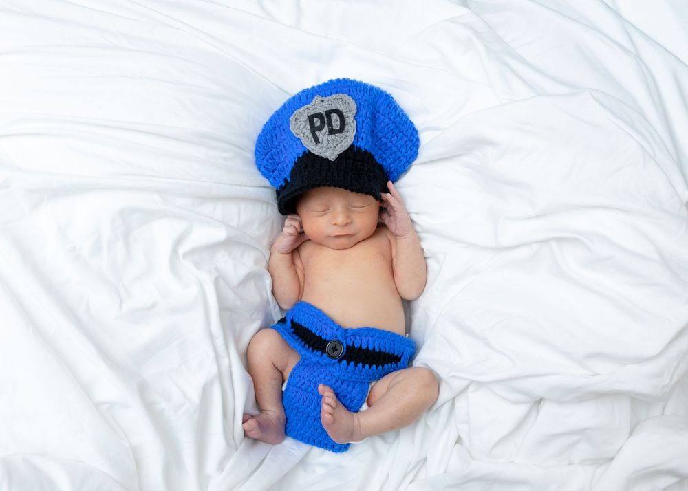 A sleeping baby wearing a blue mouse ear hat lies on white bedding next to a matching plush toy.