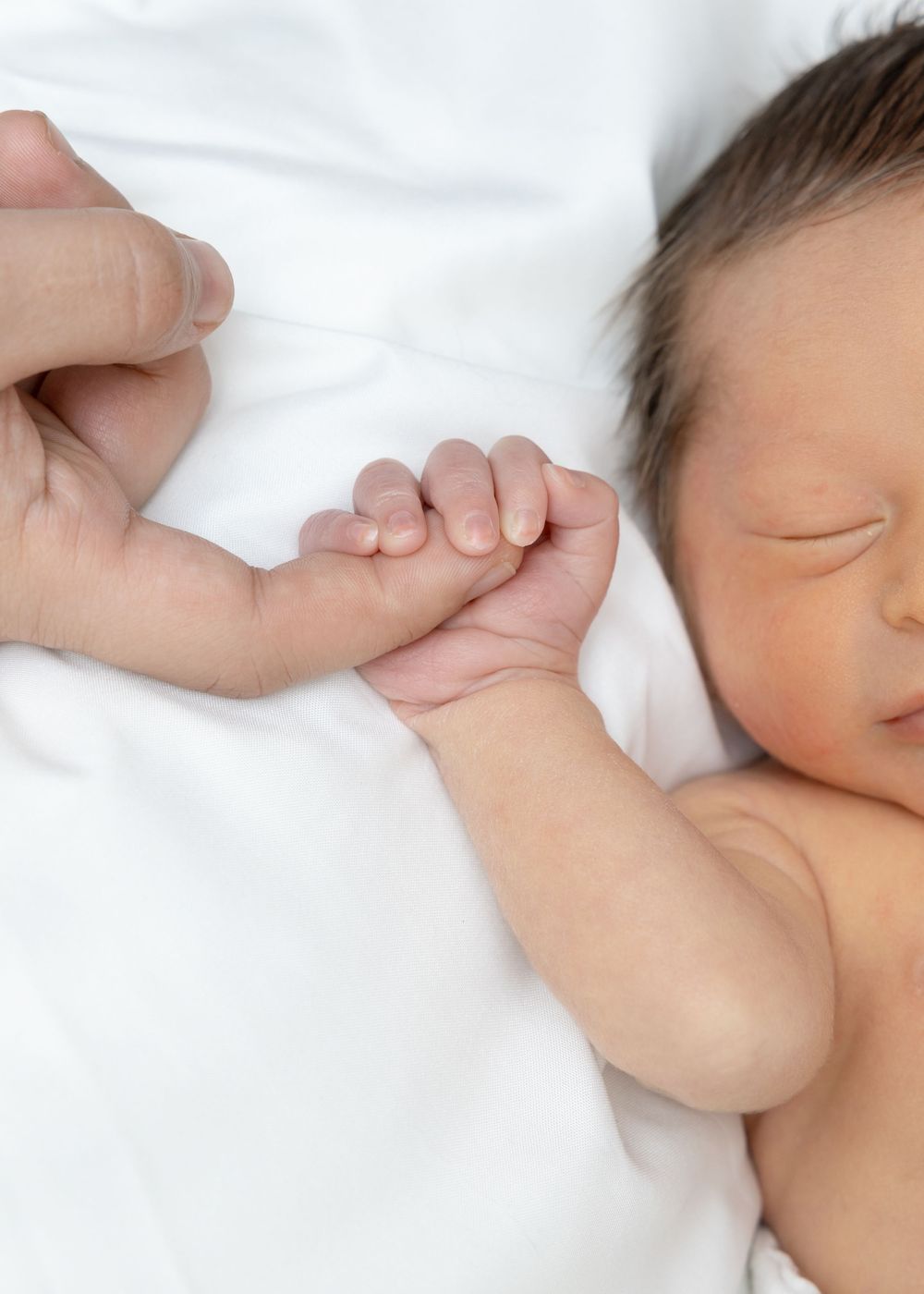 A sleeping newborn baby holds an adult finger while resting peacefully on white bedding.