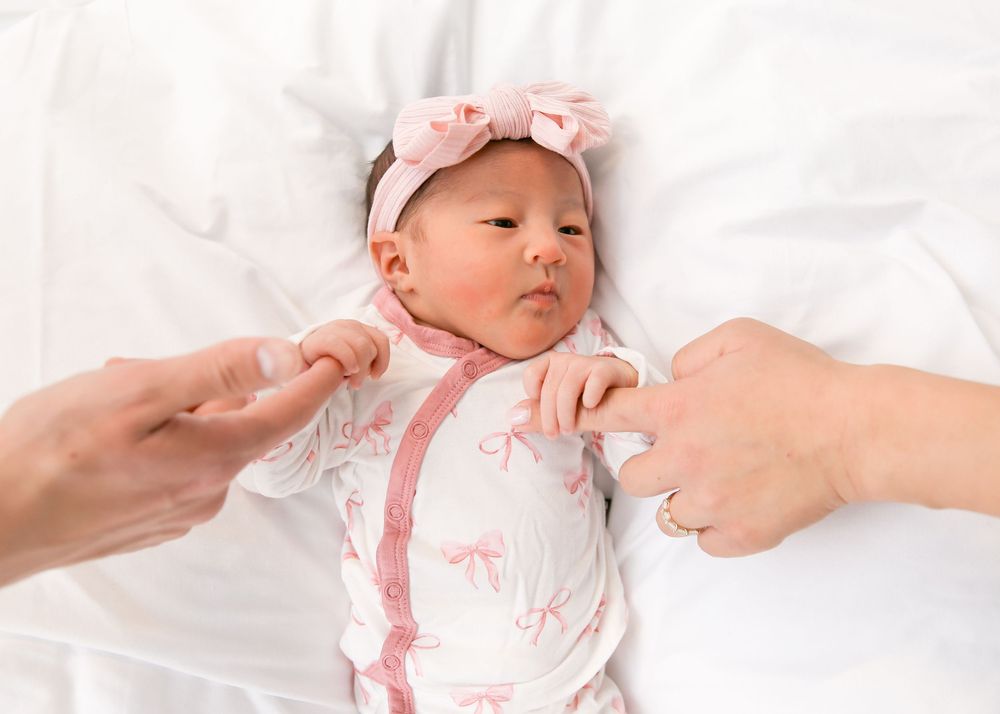 A newborn baby wearing pink and white clothing lies on white bedding while hands reach to touch them.