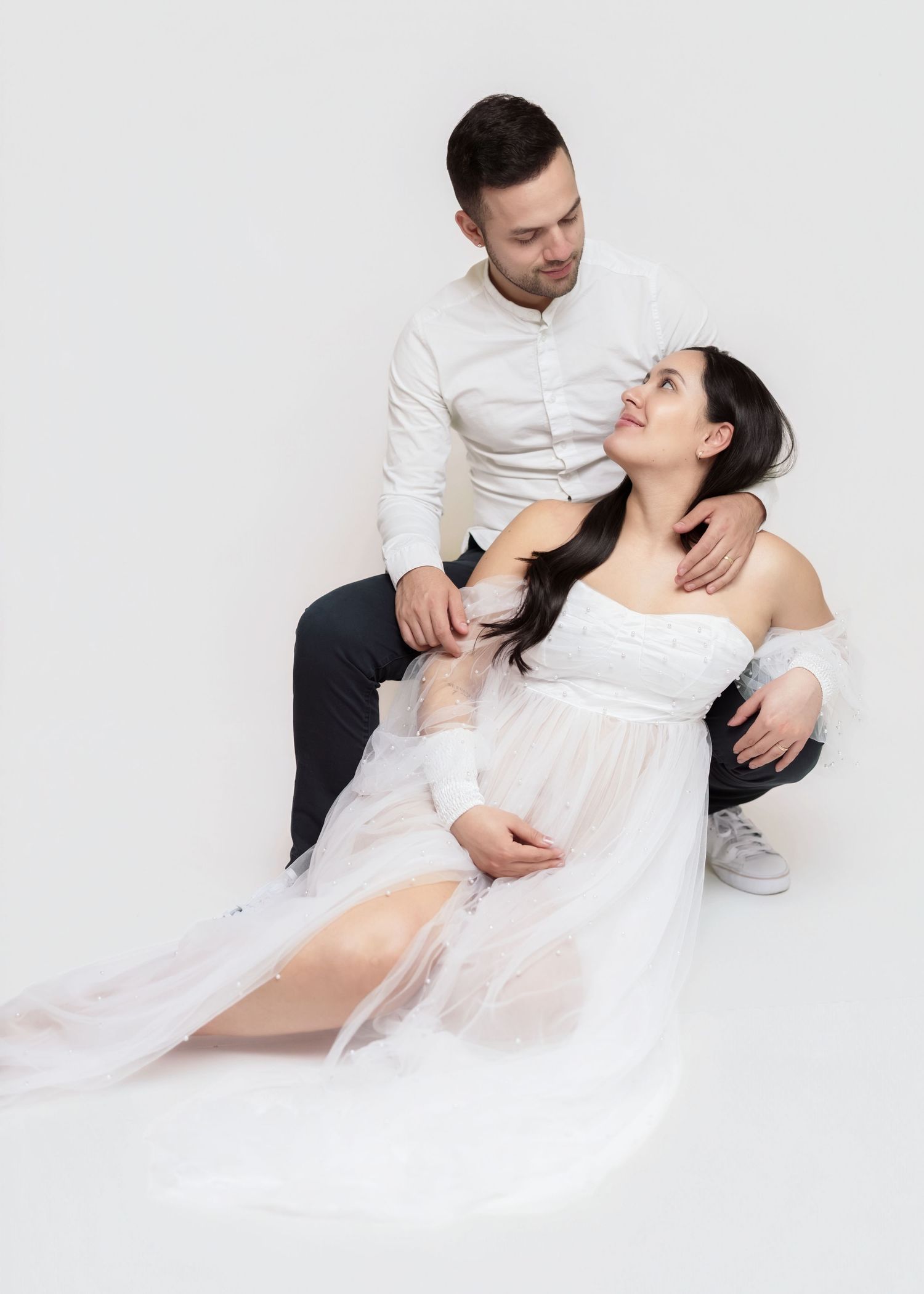 Romantic couple in white clothing poses together on light background during intimate studio photoshoot.
