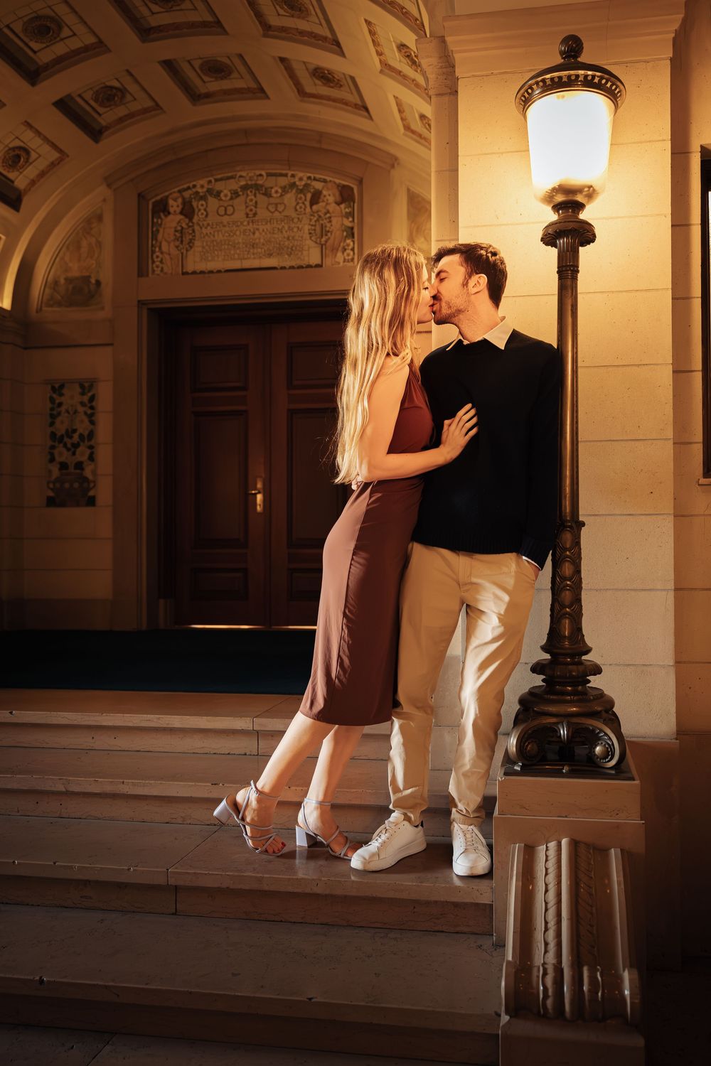A couple shares an intimate moment in a warmly lit hallway with ornate architectural details.