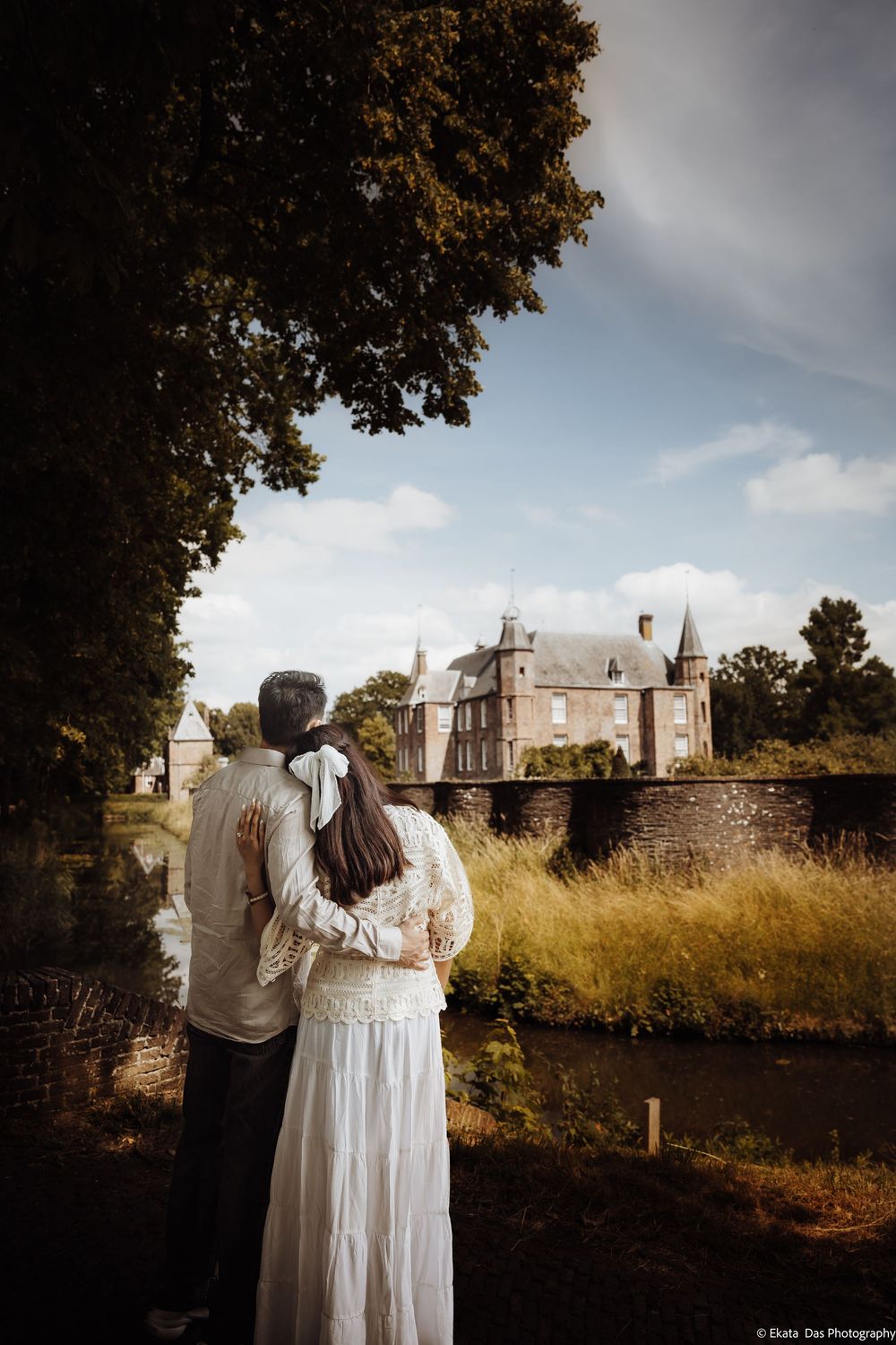Couple in wedding attire embracing while gazing at a historic castle surrounded by lush greenery and a moat.