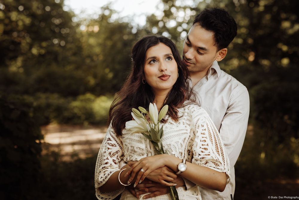 Couple embracing outdoors in lush green forest, woman holding white flowers, wearing elegant white lace dress.