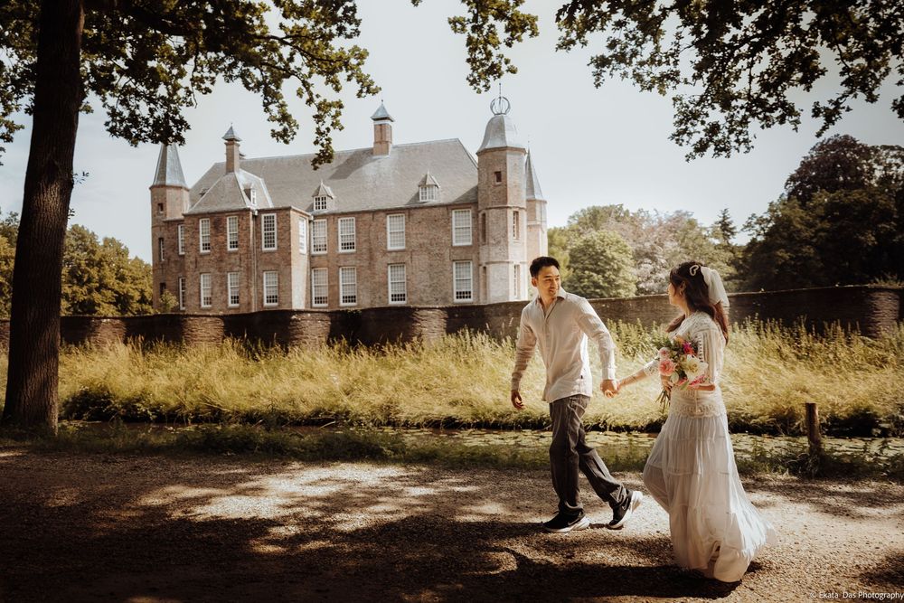 Couple holding hands walking near a historic brick castle surrounded by lush greenery on a sunny day.